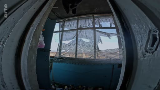 Vue en contre-plongée depuis une pièce sombre et délabrée, avec des rideaux déchirés, donnant sur un paysage extérieur aride, illustrant un contexte de conflit ou de dévastation.