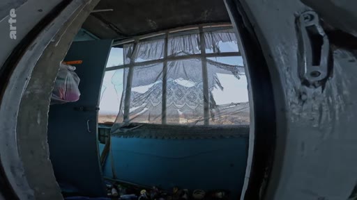 Vue en contre-plongée depuis l'intérieur d'une pièce dévastée, montrant des rideaux en dentelle déchirés et des fenêtres brisées, évoquant un contexte de conflit ou d'abandon.