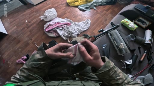 Un soldat en tenue de camouflage nettoie une arme de type fusil d'assaut avec un chiffon dans un intérieur domestique encombré, illustrant la préparation tactique ou l'entretien du matériel militaire.