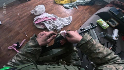 Un soldat en tenue de camouflage militaire nettoie méticuleusement son fusil d'assaut à l'intérieur d'une pièce en désordre, illustrant la préparation au combat ou l'entretien du matériel lors d'un conflit armé.