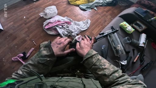 Un soldat en tenue de camouflage nettoie minutieusement son fusil d'assaut, probablement un AK, dans un intérieur encombré. Cette image illustre le quotidien et l'entretien du matériel militaire sur le terrain.
