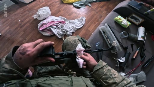 Un soldat en tenue de camouflage nettoie minutieusement le mécanisme de son fusil d'assaut à l'aide d'un chiffon, dans une pièce encombrée. Cette image illustre la maintenance du matériel militaire en zone de conflit.