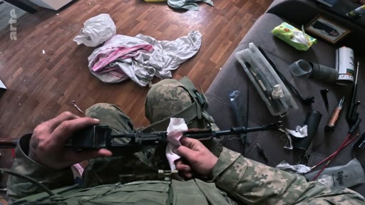 Un soldat en tenue de camouflage nettoie méticuleusement son fusil d'assaut avec un chiffon, vu à la première personne dans une pièce en désordre. Cette image illustre la préparation et l'entretien du matériel militaire sur le terrain.
