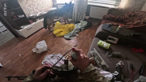 Un soldat en tenue de camouflage nettoie son fusil d'assaut à l'intérieur d'une habitation lors d'une opération militaire. Un chien de type berger allemand se tient à proximité dans cette pièce au décor domestique improvisé.