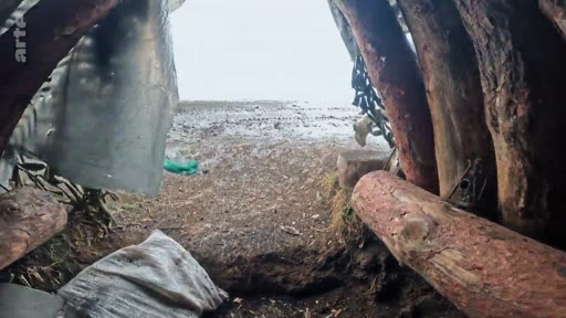 Vue en perspective depuis l'intérieur d'une tranchée militaire fortifiée avec des parois en rondins de bois, donnant sur un terrain extérieur enneigé ou boueux. Cette image illustre les conditions de vie et de protection des soldats sur le front.