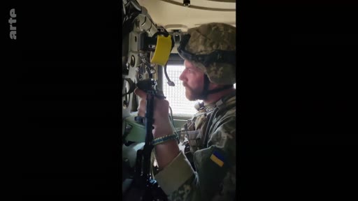 Un soldat ukrainien portant un casque et un uniforme avec le drapeau de l'Ukraine est assis à l'intérieur d'un véhicule blindé, manipulant un équipement. Cette image illustre le quotidien des troupes ukrainiennes sur le front.
