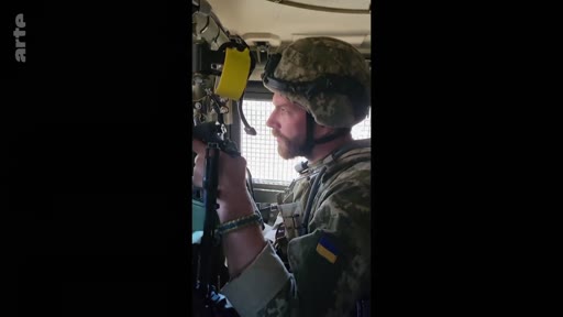 Un soldat ukrainien portant un casque et un uniforme avec le drapeau de l'Ukraine est filmé à l'intérieur d'un véhicule blindé. Cette image illustre le quotidien des troupes ukrainiennes sur le front lors du conflit en Ukraine.