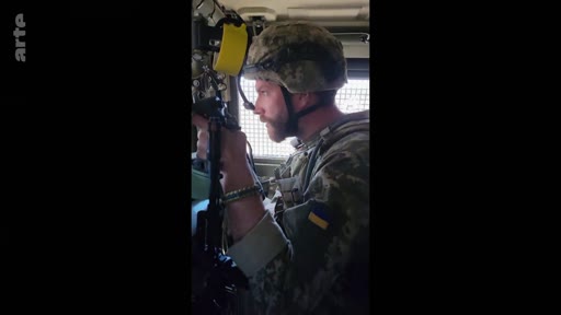 Un soldat ukrainien portant un casque et un uniforme avec le drapeau de l'Ukraine est filmé à l'intérieur d'un véhicule militaire. Cette image illustre le quotidien des troupes ukrainiennes sur le front.