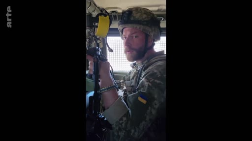 Un soldat ukrainien portant un casque et un uniforme avec le drapeau de l'Ukraine est filmé à l'intérieur d'un véhicule blindé. Cette image illustre le quotidien des militaires sur le front lors du conflit en Ukraine.