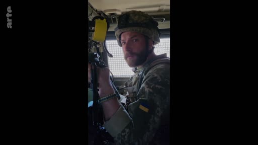 Un soldat ukrainien portant un casque et un uniforme de camouflage avec un écusson aux couleurs de l'Ukraine est filmé à l'intérieur d'un véhicule blindé. Cette image illustre le quotidien des militaires sur le front lors du conflit en Ukraine.