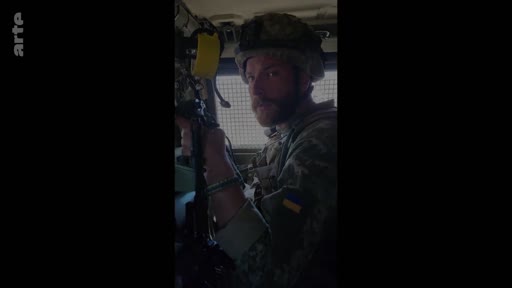 Un soldat ukrainien portant un casque et un uniforme avec le drapeau de l'Ukraine est filmé à l'intérieur d'un véhicule militaire. Cette image illustre le quotidien des troupes ukrainiennes sur le front.