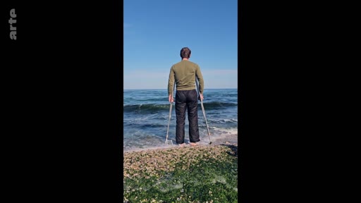 Un homme vu de dos, utilisant des béquilles pour se tenir debout au bord de l'eau sur une plage, illustrant un moment de solitude et de résilience face à l'océan.