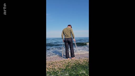 Un homme blessé ou en convalescence se tient debout sur le rivage d'une plage, s'appuyant sur des béquilles tout en regardant l'océan. Cette image illustre la résilience et la recherche de calme dans la nature.