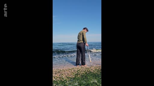 Un homme blessé, utilisant une béquille pour se déplacer, se tient au bord de l'eau sur une plage ensoleillée. Cette image illustre la résilience et la recherche de calme dans la nature malgré un handicap physique.