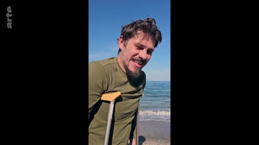Un homme souriant, s'appuyant sur des béquilles, profite d'un moment de détente sur une plage ensoleillée. Cette image illustre la résilience et la recherche de bien-être en extérieur.