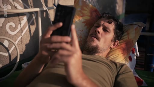 Un homme barbu, allongé sur un lit avec un oreiller à motifs, consulte son téléphone portable, illustrant le quotidien et la solitude d'un soldat ou d'une personne marquée par le conflit.