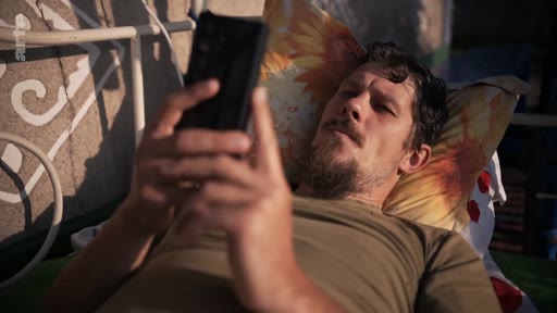 Un homme barbu, allongé sur un lit avec un oreiller à motifs, regarde attentivement l'écran de son smartphone. Cette scène illustre le quotidien et la solitude d'un individu, potentiellement en lien avec le traumatisme ou le repos après un conflit.