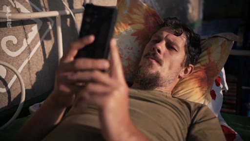 Un homme barbu, allongé sur un lit, consulte son téléphone portable, illustrant le quotidien et la solitude d'un soldat ou d'une personne marquée par le conflit.