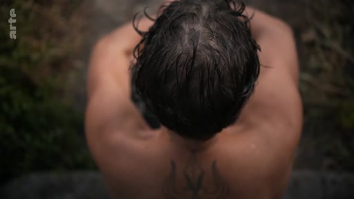 Vue en plongée d'un homme torse nu portant un tatouage distinctif dans le dos, illustrant le thème des traumatismes psychologiques liés à la guerre.