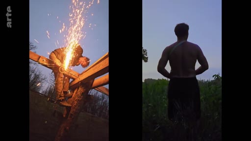 Un homme utilise une meuleuse pour travailler sur une structure en bois dans le cadre de la construction du centre Goloka, tandis qu'un autre homme torse nu observe les environs au crépuscule.