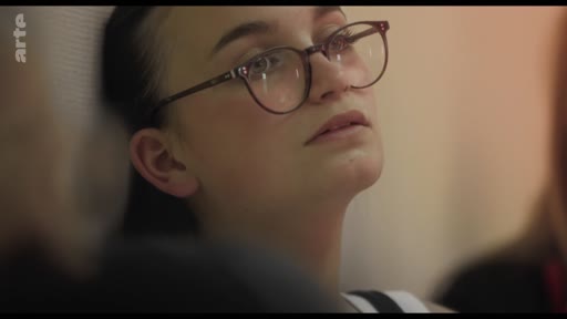 Une jeune femme aux cheveux bruns et portant des lunettes est filmée en gros plan alors qu'elle écoute une intervention lors d'une réunion de groupe.
