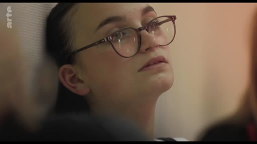 Une jeune femme aux cheveux attachés et portant des lunettes est filmée en gros plan alors qu'elle écoute une intervention lors d'une réunion de groupe.