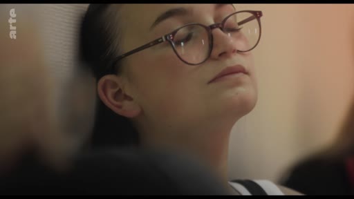 Une jeune femme aux cheveux attachés et portant des lunettes est filmée en gros plan alors qu'elle semble écouter une intervention lors d'une réunion ou d'un atelier.