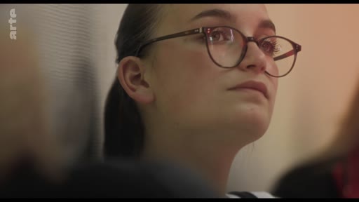 Une jeune femme avec des lunettes, vue en gros plan, écoute attentivement lors d'une réunion ou d'une séance de travail. Elle semble concentrée sur les propos tenus par le directeur ou l'intervenant hors champ.