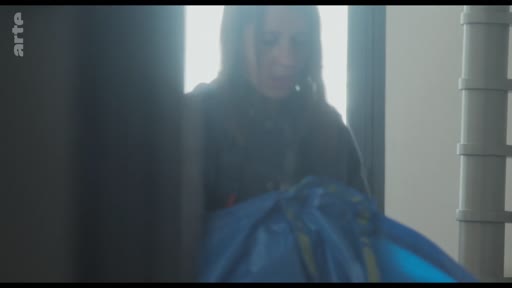 Une femme, vue en plan moyen, range des objets dans un grand sac de transport bleu, probablement lors d'un déménagement ou d'un départ.