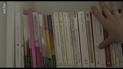 Une main saisit un exemplaire du livre 'Alice au pays des merveilles' de Lewis Carroll dans une bibliothèque personnelle. On aperçoit également des ouvrages d'Agota Kristof et Ian McEwan sur l'étagère.