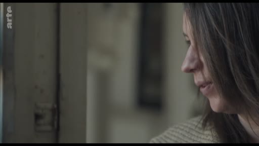 Une femme aux cheveux bruns est filmée en gros plan de profil, regardant pensivement par une fenêtre dans une scène silencieuse.