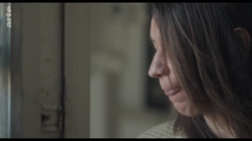 Une femme aux cheveux bruns est filmée en gros plan, regardant par la fenêtre avec une expression pensive et mélancolique. L'image capture un moment d'introspection silencieuse dans un cadre domestique.