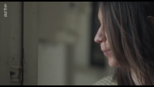 Une femme aux cheveux bruns est filmée en gros plan de profil alors qu'elle regarde à travers l'entrebâillement d'une porte dans une scène silencieuse.