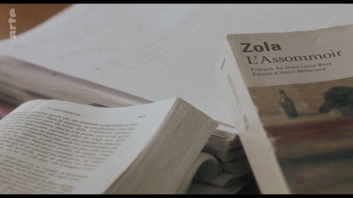 Gros plan sur une édition du roman L'Assommoir d'Émile Zola, posée sur une table aux côtés d'autres documents, illustrant une analyse littéraire de l'œuvre naturaliste.