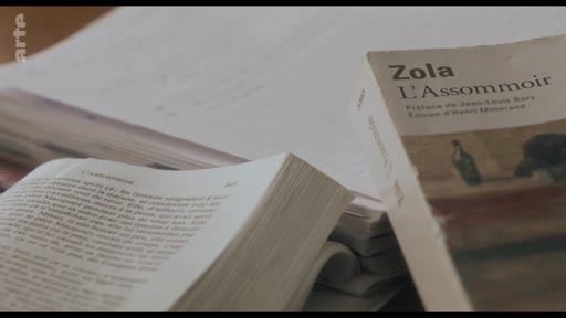 Gros plan sur une édition du roman naturaliste L'Assommoir d'Émile Zola, posée sur une pile de documents et de livres, illustrant une analyse littéraire de l'œuvre.