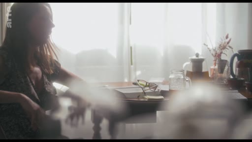 Une femme est filmée en plan moyen dans une pièce baignée de lumière, assise à une table avec des objets personnels, lors d'une interview pour le programme 'Crafting the Character'.