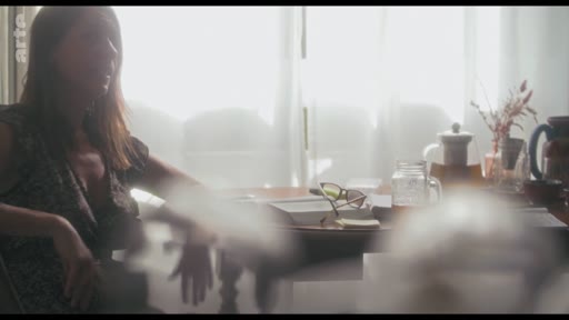 Une femme est filmée en plan moyen lors d'une interview, assise à une table avec des objets personnels, dans une ambiance lumineuse et douce.