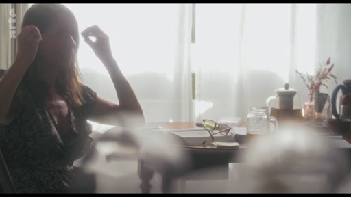 Une femme est filmée en contre-jour alors qu'elle est assise à une table, se touchant les cheveux dans un moment de réflexion ou de préparation. La scène, baignée par la lumière d'une fenêtre, évoque une atmosphère intime et personnelle lors d'une interview.
