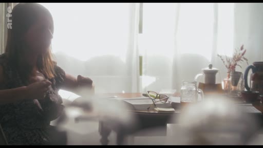 Une femme est assise à une table dans une pièce baignée de lumière, avec des objets personnels posés devant elle, dans le cadre d'une interview sur la création de personnage.