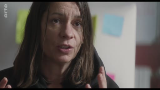 Une femme s'exprime face caméra lors d'une interview, avec des post-its colorés visibles en arrière-plan. La scène semble se dérouler dans un cadre de travail ou de réflexion.