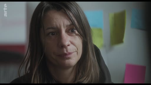 Une actrice, filmée en gros plan, écoute attentivement les instructions lors d'une séance de travail ou d'une discussion sur le plateau de tournage. Des post-its colorés sont visibles en arrière-plan sur un mur.