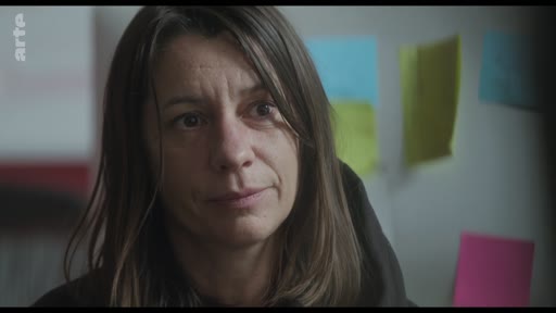 Une femme, probablement une actrice ou une réalisatrice, est filmée en gros plan lors d'une discussion sur le plateau de tournage. En arrière-plan, des post-its colorés sont visibles sur un mur.