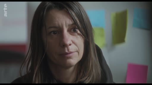 Une femme, probablement une actrice ou une réalisatrice, est filmée en gros plan lors d'une discussion sur le plateau de tournage. Elle semble attentive et concentrée, avec des notes adhésives colorées visibles en arrière-plan.