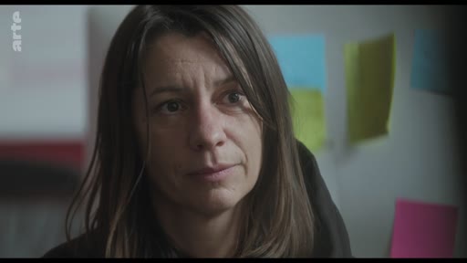 Une femme, probablement une actrice ou une réalisatrice, est filmée en gros plan lors d'une discussion sur le plateau de tournage. En arrière-plan, des post-its colorés sont visibles sur un mur, suggérant un environnement de travail créatif ou de préparation.