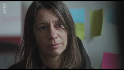 Une femme, probablement une actrice, écoute attentivement lors d'une discussion sur le plateau de tournage. Elle est filmée en gros plan avec des post-its colorés visibles en arrière-plan.