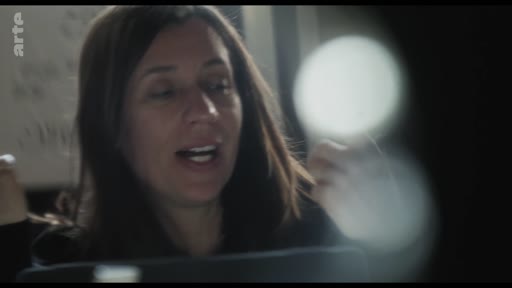 Une femme s'exprime lors d'une réunion de travail dans un espace de production, probablement en train de faire un débriefing.