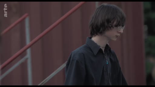 Un jeune homme aux cheveux bruns, vêtu d'une chemise noire, est filmé en plan moyen lors d'une discussion de groupe au théâtre.
