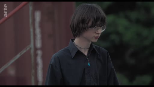 Un jeune homme aux cheveux bruns et portant des lunettes, vêtu d'une chemise noire, est filmé en plan moyen lors d'une discussion au théâtre.