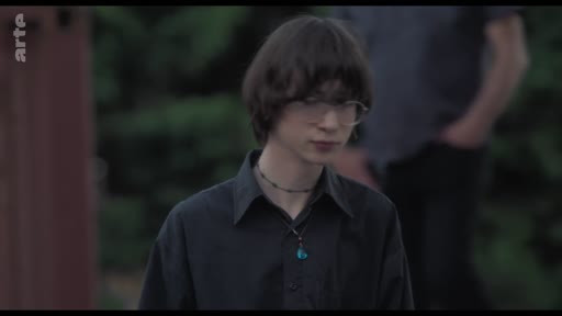 Un jeune homme aux cheveux bruns, portant des lunettes et une chemise noire, participe à une discussion de groupe dans un théâtre.
