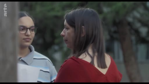 Deux femmes, dont une portant des lunettes et un polo rayé, discutent lors d'une répétition de théâtre en extérieur. La scène montre une interaction entre les deux protagonistes dans un cadre informel.